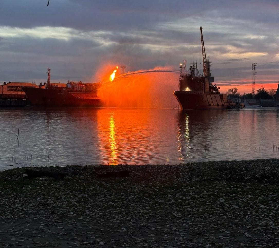 A firefighting vessel sprays water on a burning ship at dusk, reflecting orange flames.