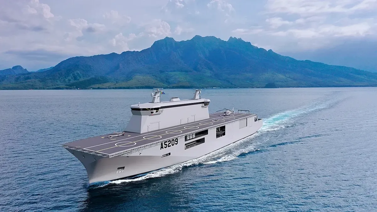 Large grey naval ship with flight deck for drones cruising near mountains.