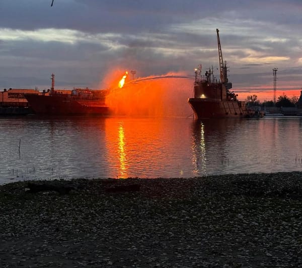 A firefighting vessel sprays water on a burning ship at dusk, reflecting orange flames.